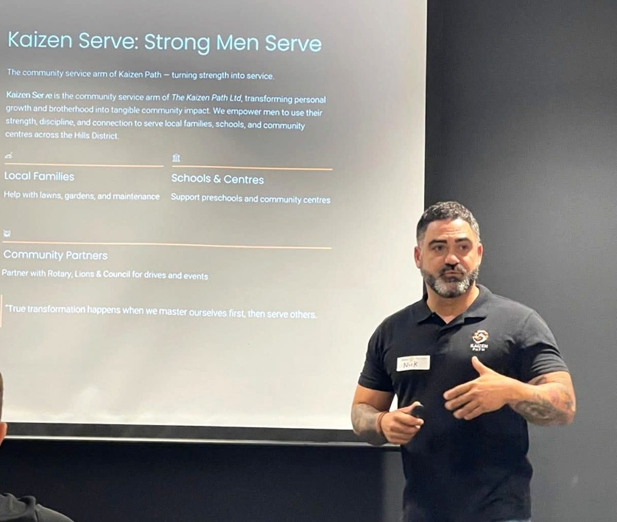 Nick Stamatis speaking to a seated audience beside a Kaizen Serve presentation slide